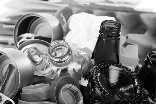 A close-up view of a collection of various discarded packaging and waste materials arranged on a surface, including crumpled paper, empty glass and plastic bottles with open caps, aluminum and plastic drink cans, and a black glass bottle. Surrounding the waste are plastic lids and a metallic object resembling a decorative bowl or container with a beaded edge. The background is blurred but suggests an indoor environment, possibly a storage area or space designated for rubbish. This scene illustrates general waste typical of rubbish removal services in Forest Gate, with materials that require proper collection and disposal by a professional waste management provider such as Business Waste Removal Forest Gate.