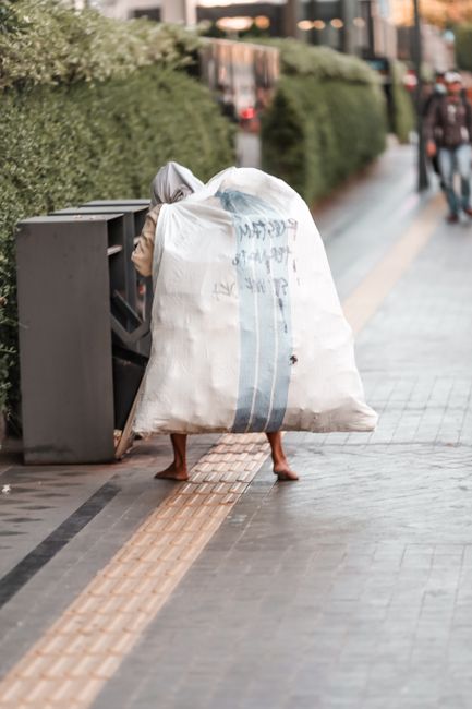 From an overhead perspective, a waste collection worker dressed in an orange high-visibility jacket with reflective yellow-green stripes and matching orange cap is seen standing on a paved sidewalk ne