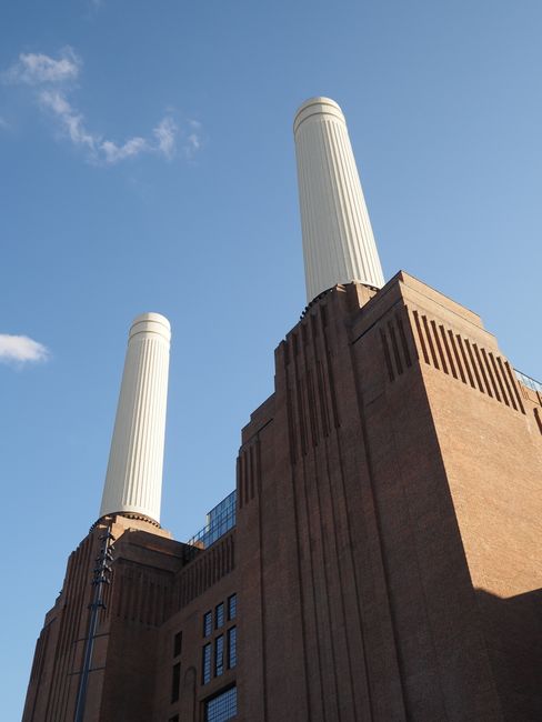 A low-angle view of a historic brick chimney structure with two tall, cylindrical white flues extending into a clear blue sky. The brickwork features vertical ridges and decorative sections, with some
